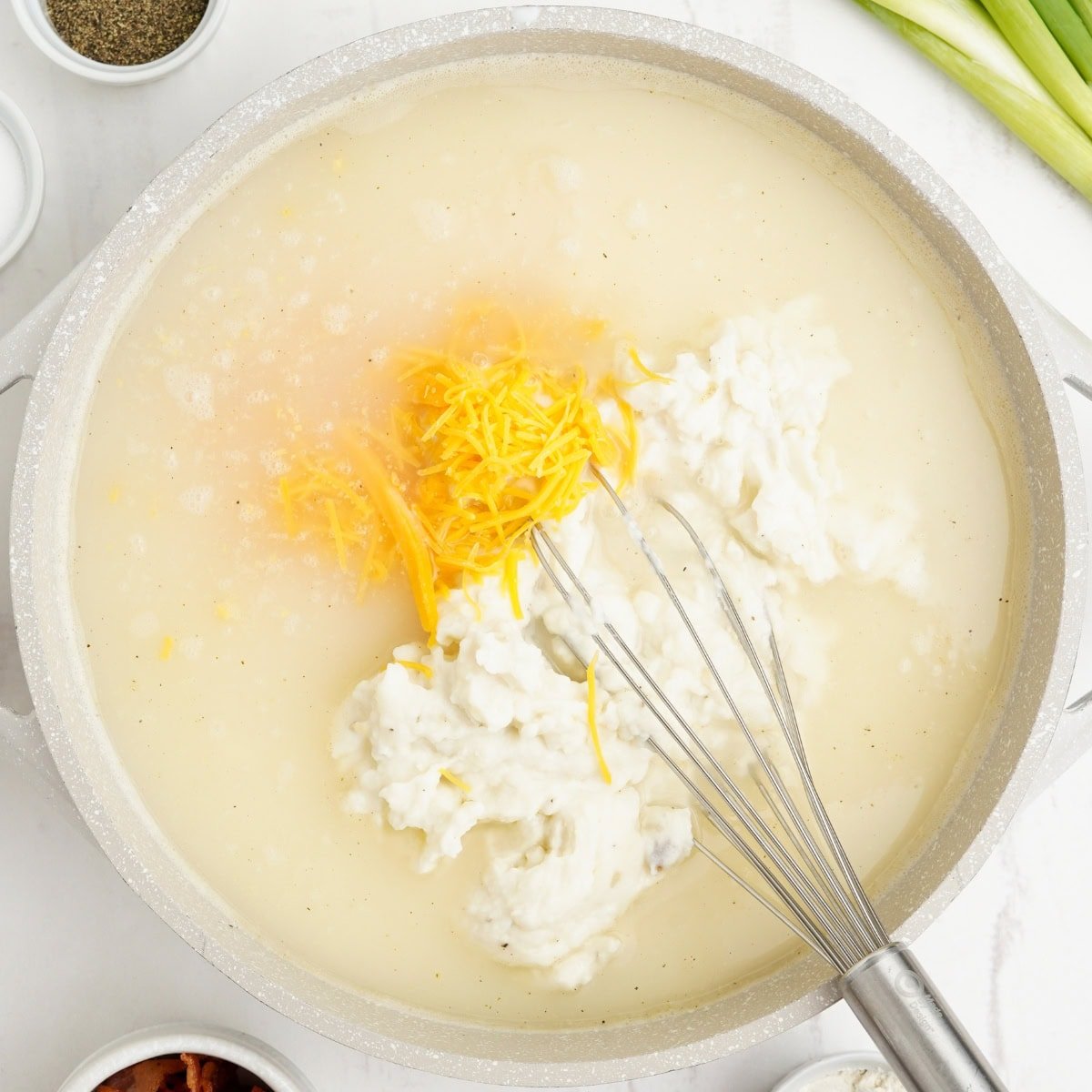 A whisk rests on the side of a pot holding broth, mashed potatoes, and cheese.