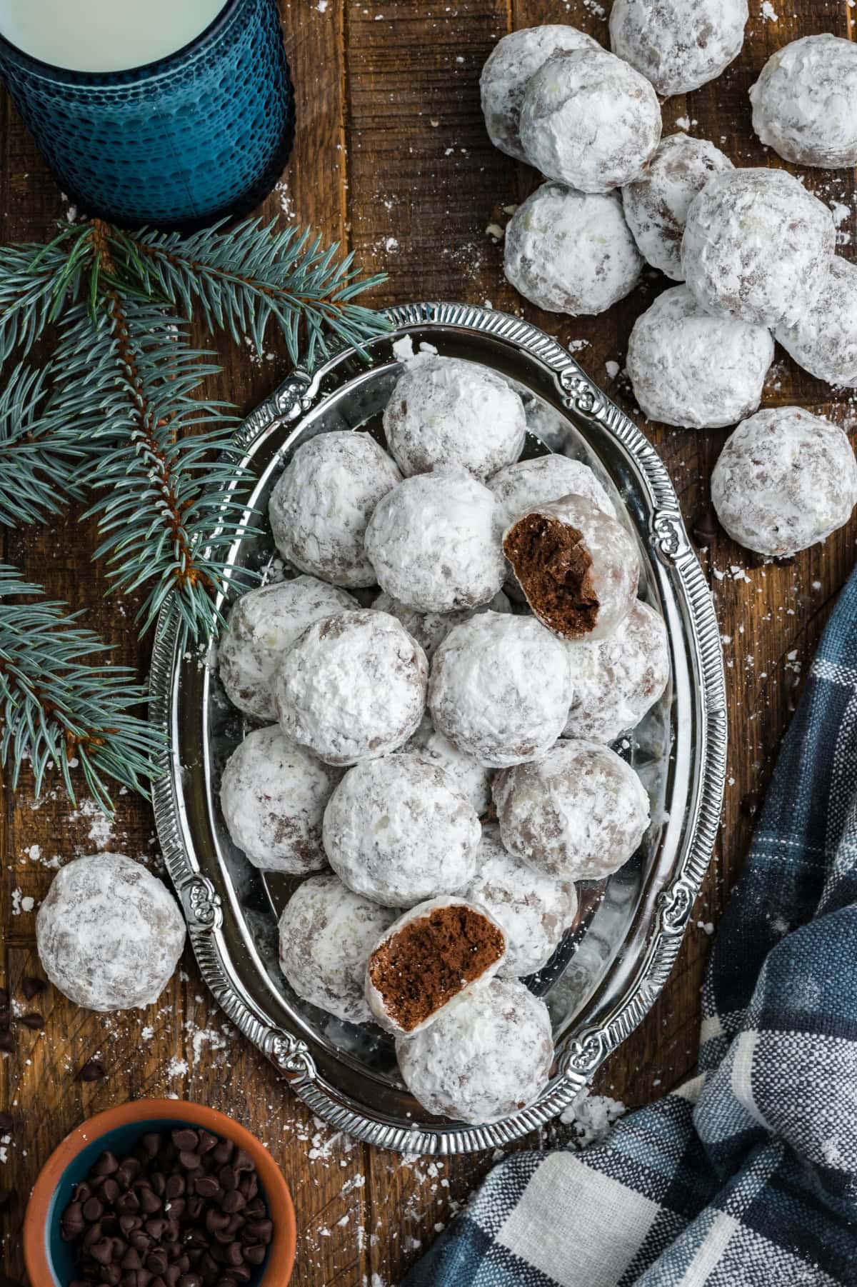 A silver platter of chocolate snowball cookies next to sprigs of evergreen and a plaid dishtowel.