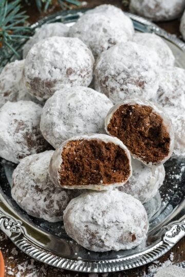 A silver plate arranged with chocolate snowball cookies. One of the cookies has been broken in half.