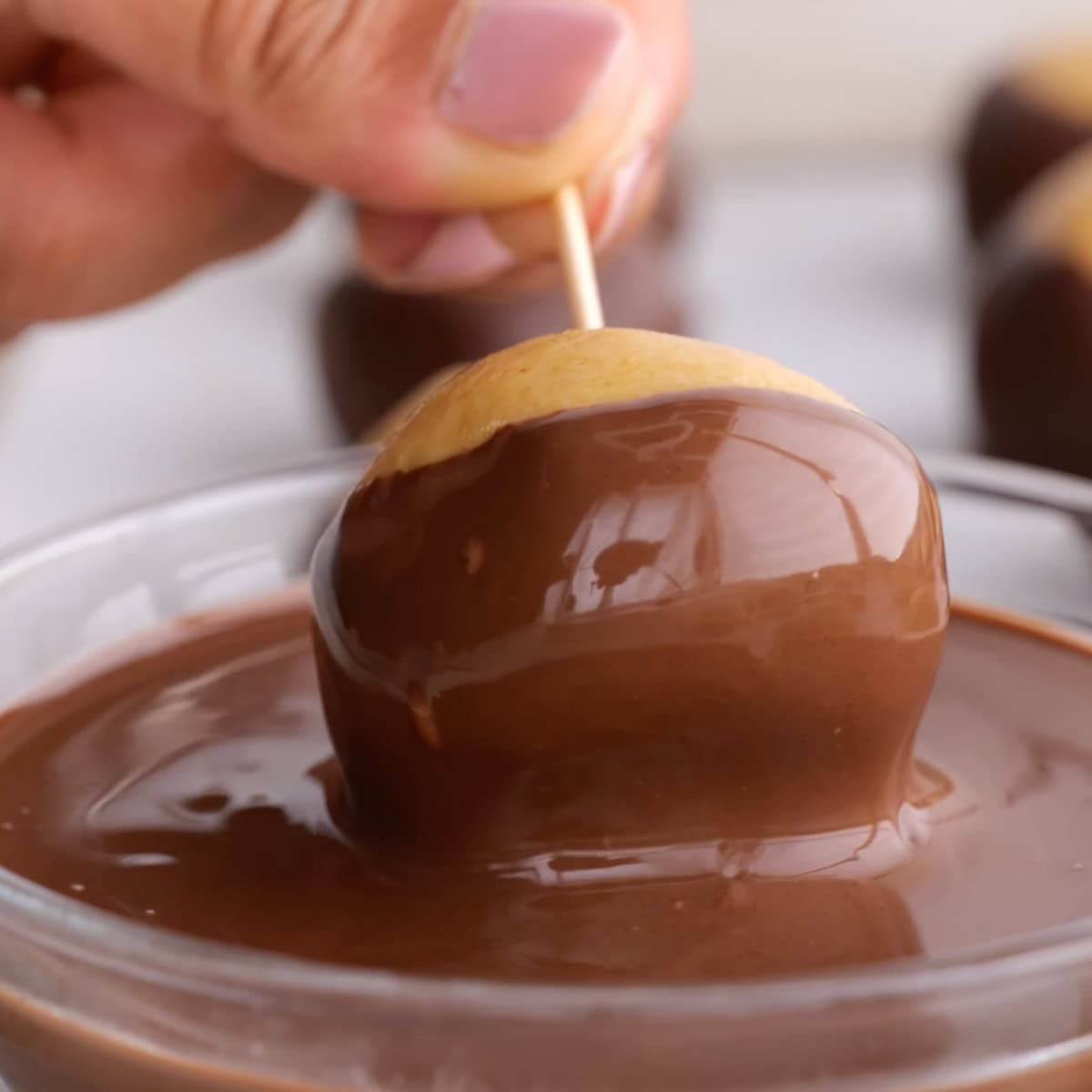 A hand uses a toothpick to dip a peanut butter ball in melted chocolate.