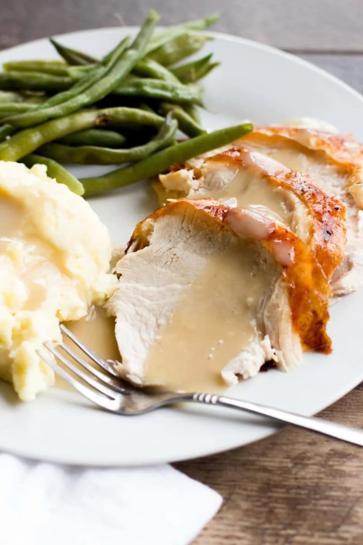A plate of sliced turkey, mashed potatoes, and green beans. The turkey and potatoes are topped with gravy.