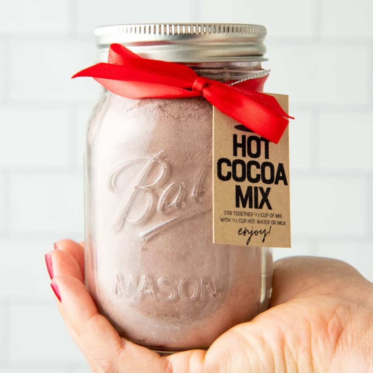 A hand holds up a jar of hot chocolate mix tied with a red ribbon and a tag.