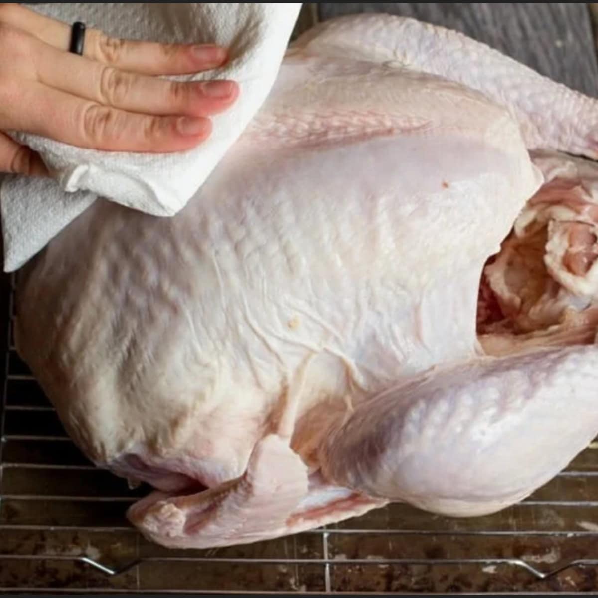 A hand uses a paper towel to pat a raw turkey dry.