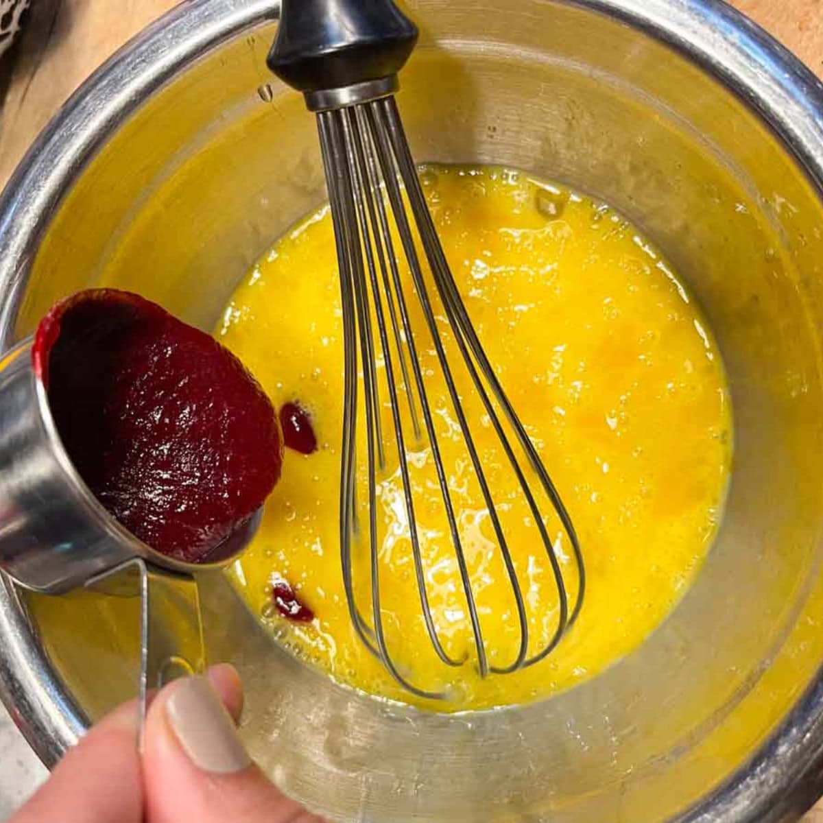 A whisk in a bowl of eggs, while a hand pours some hot cranberry filling into the bowl to temper the eggs.