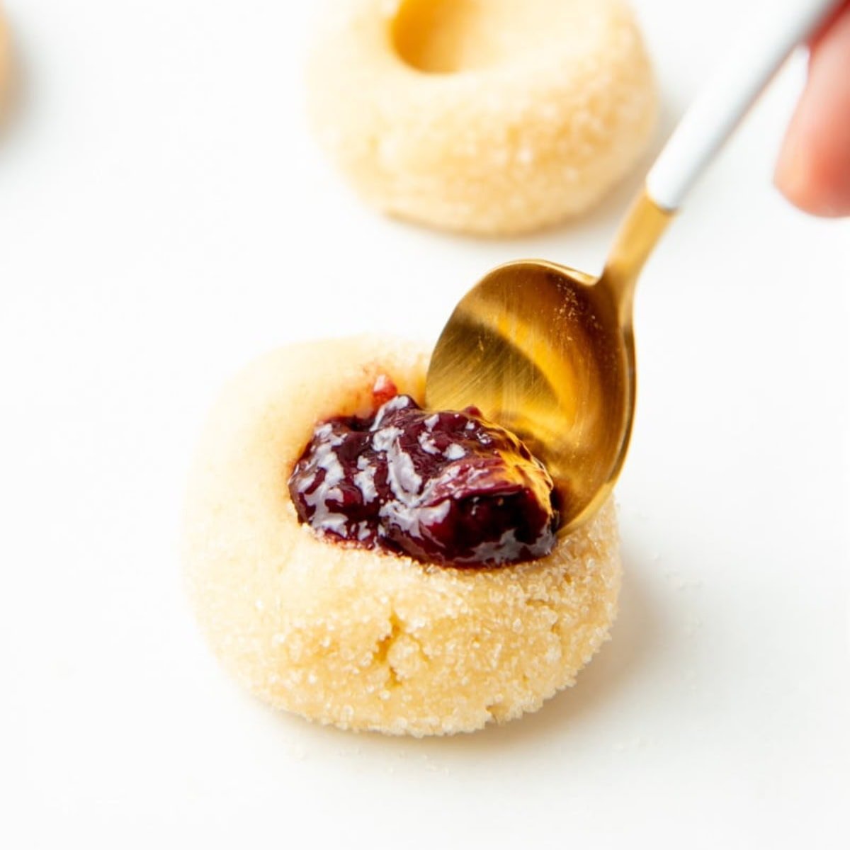 A white and gold spoon puts raspberry jam into a thumbprint cookie dough ball.