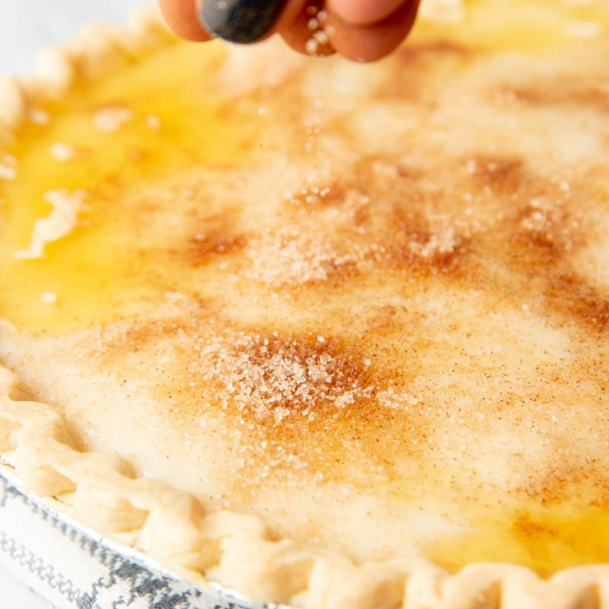 A hand sprinkles spiced sugar over the top of a sugar cream pie before baking.