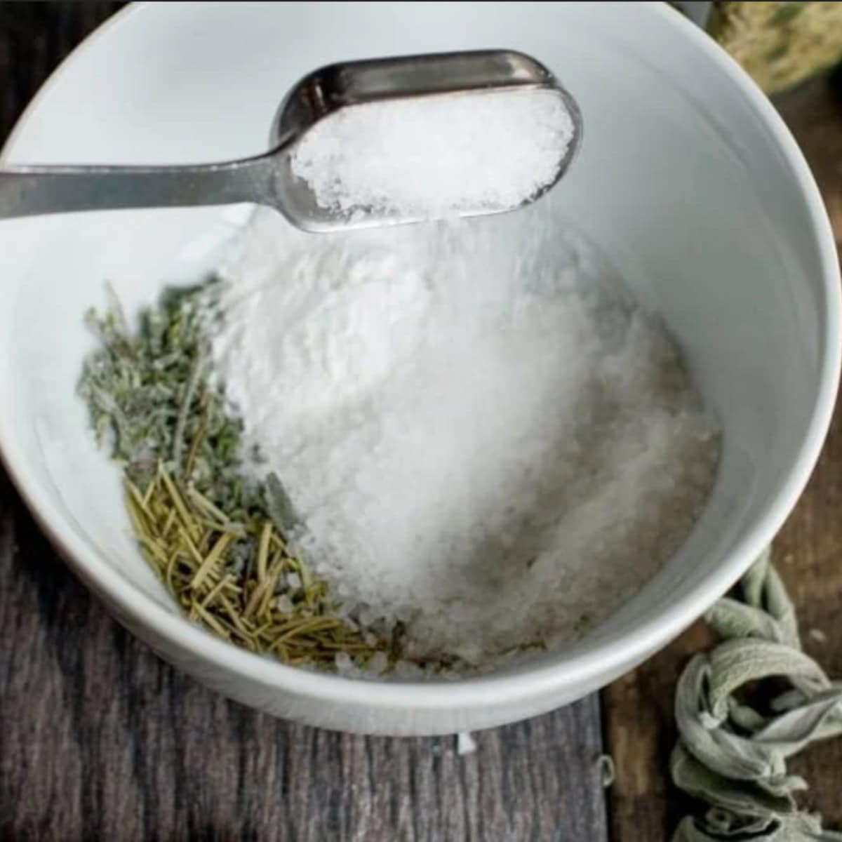 A metal measuring spoon adds salt to a bowl of seasoning and herbs.