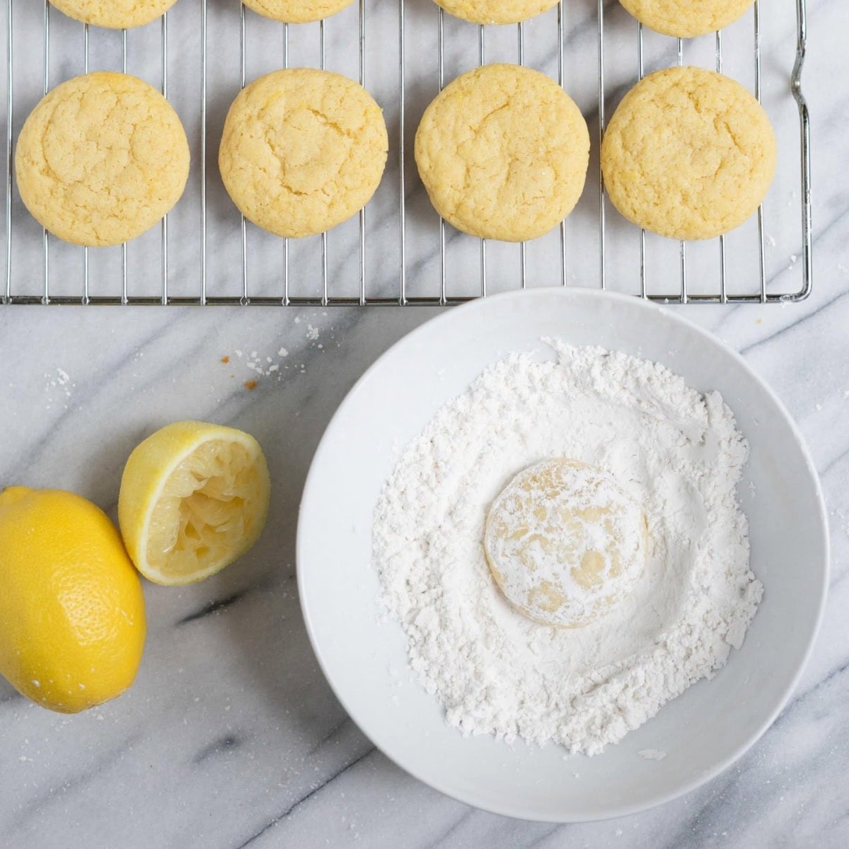 Lemon crinkle cookies on a cooling rack, with one in a bowl of powdered sugar.