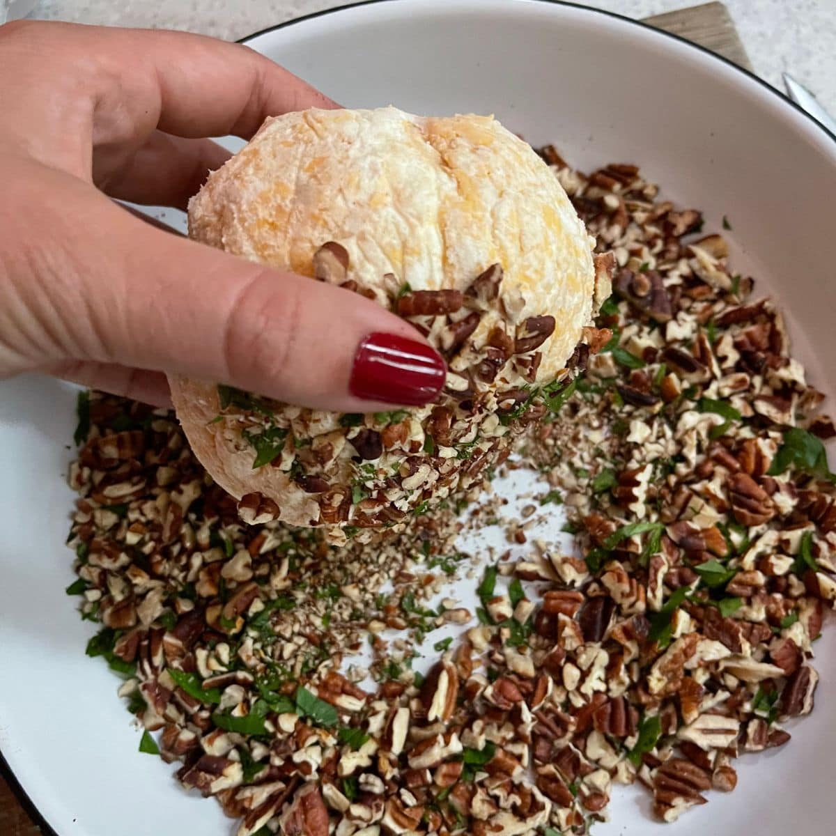 A hand rolls a cheese ball in a plate of pecans and parsley.