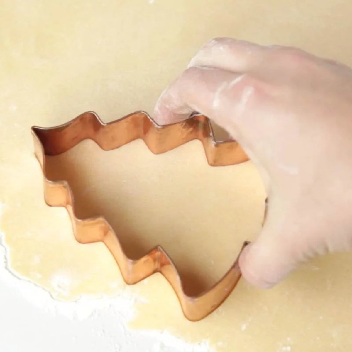 A hand uses a copper cookie cutter to cut out a tree-shaped sugar cookie.