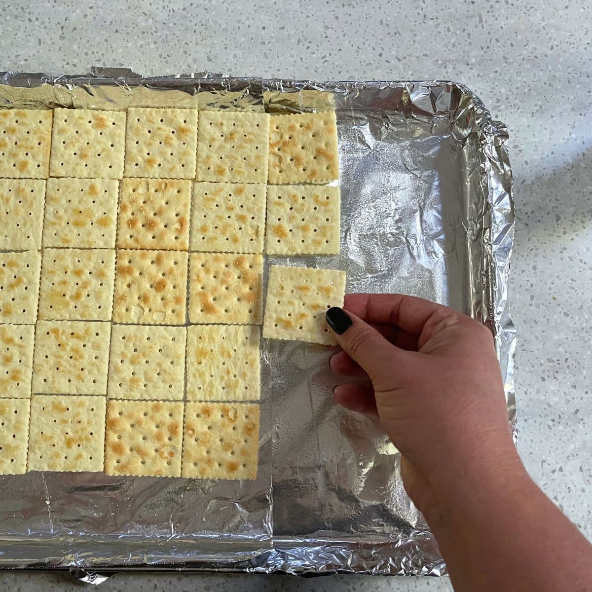 A hand arranges saltine crackers in a tight single layer.