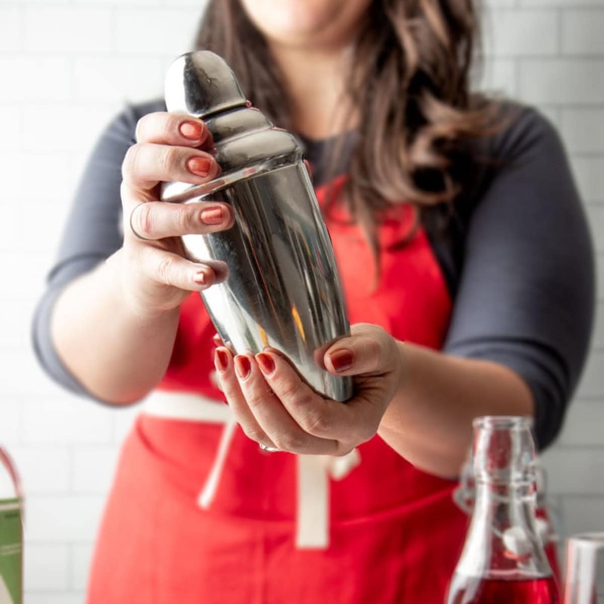 A brunette woman in a red apron shakes a silver cocktail shaker.