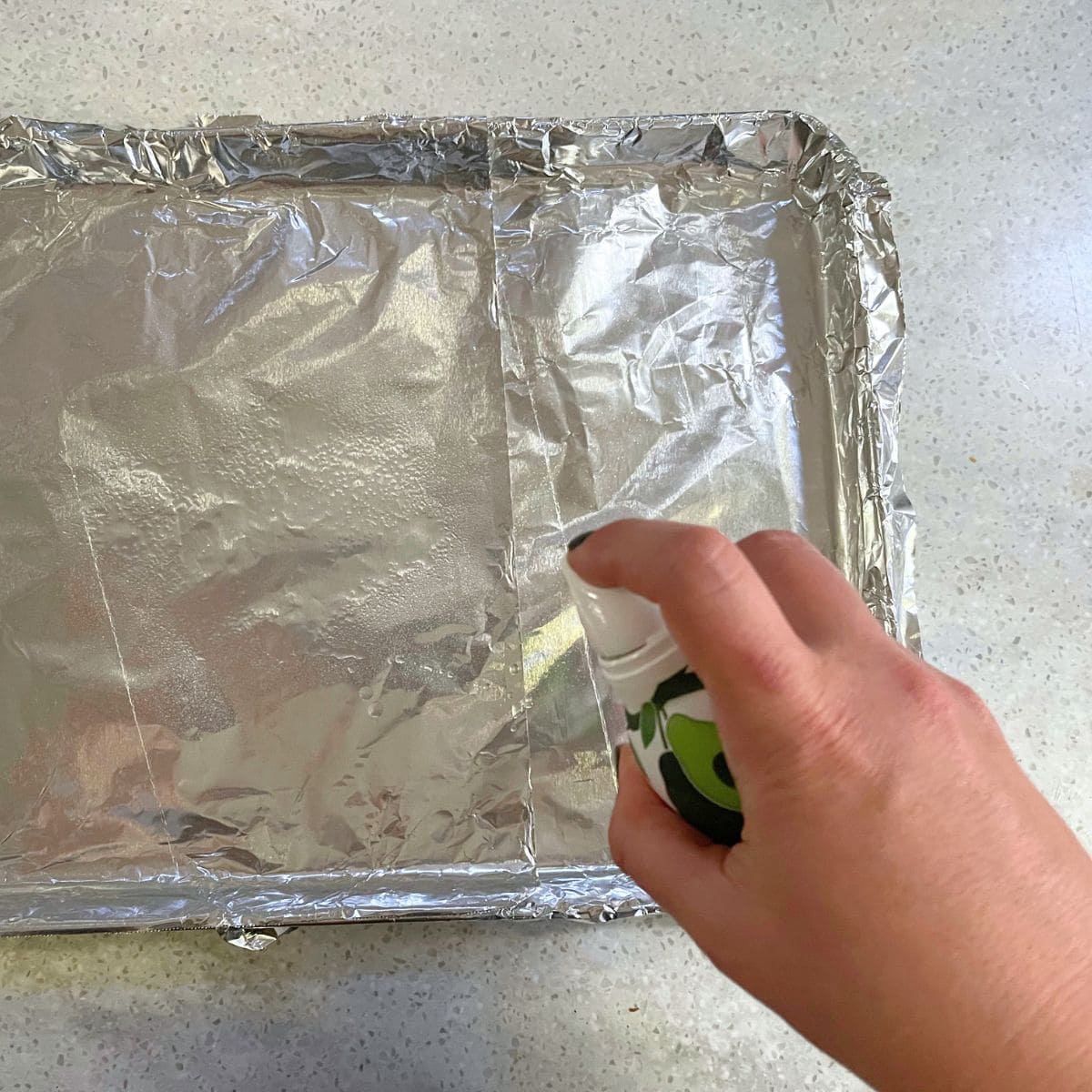 A hand sprays a foil-lined baking sheet.