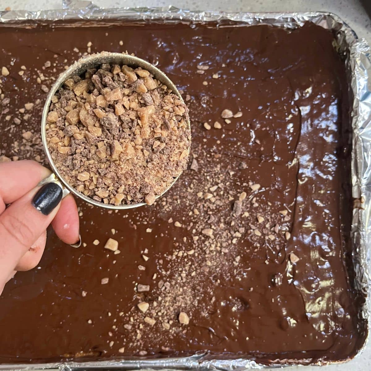 A hand sprinkles toffee bits over saltine cracker toffee.