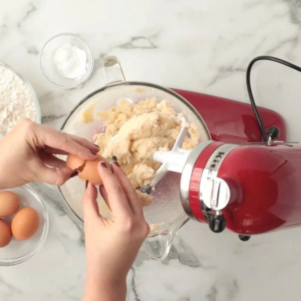 Hands add a cracked egg to the dough in a red stand mixer.