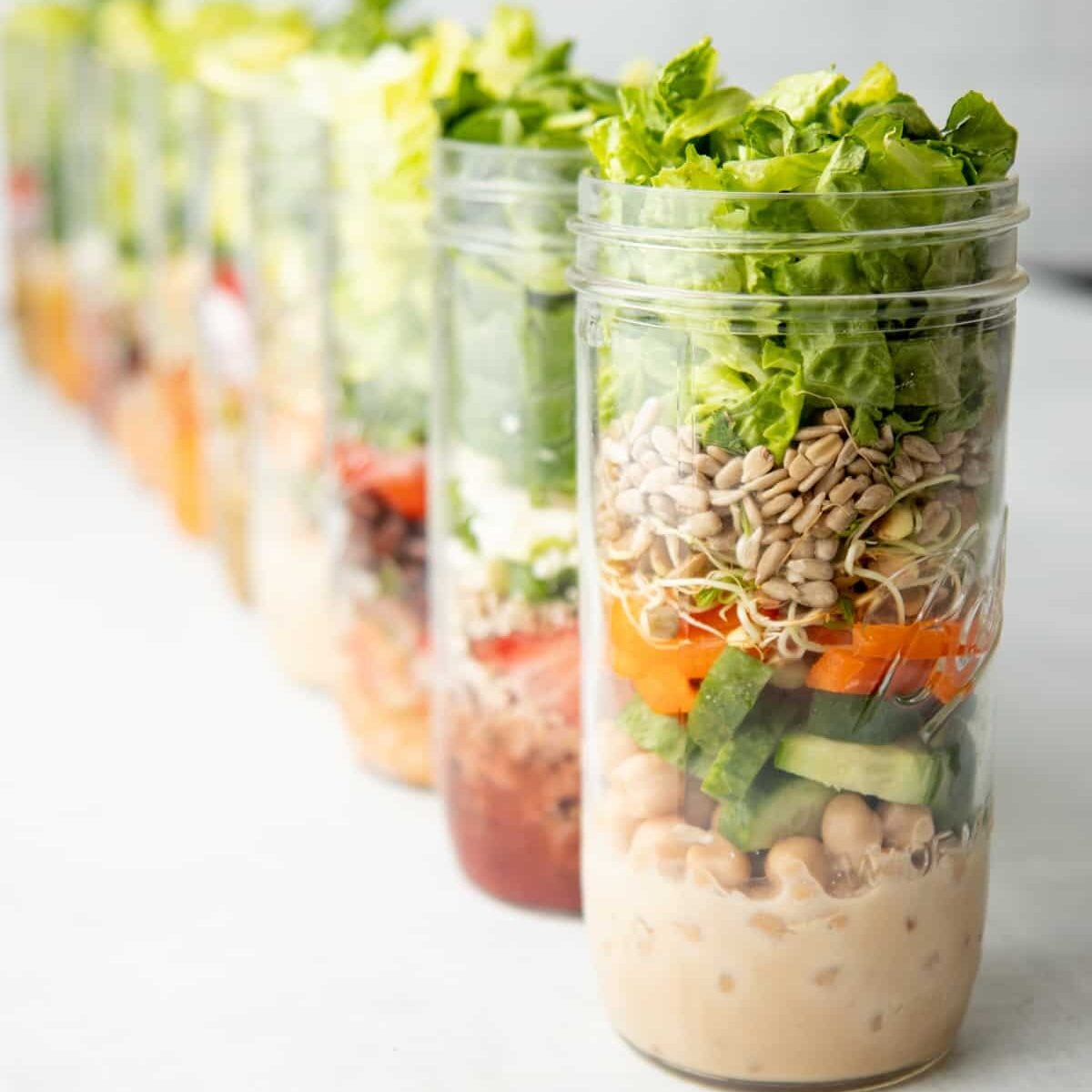 Ten tall mason jars are layered with salad ingredients.