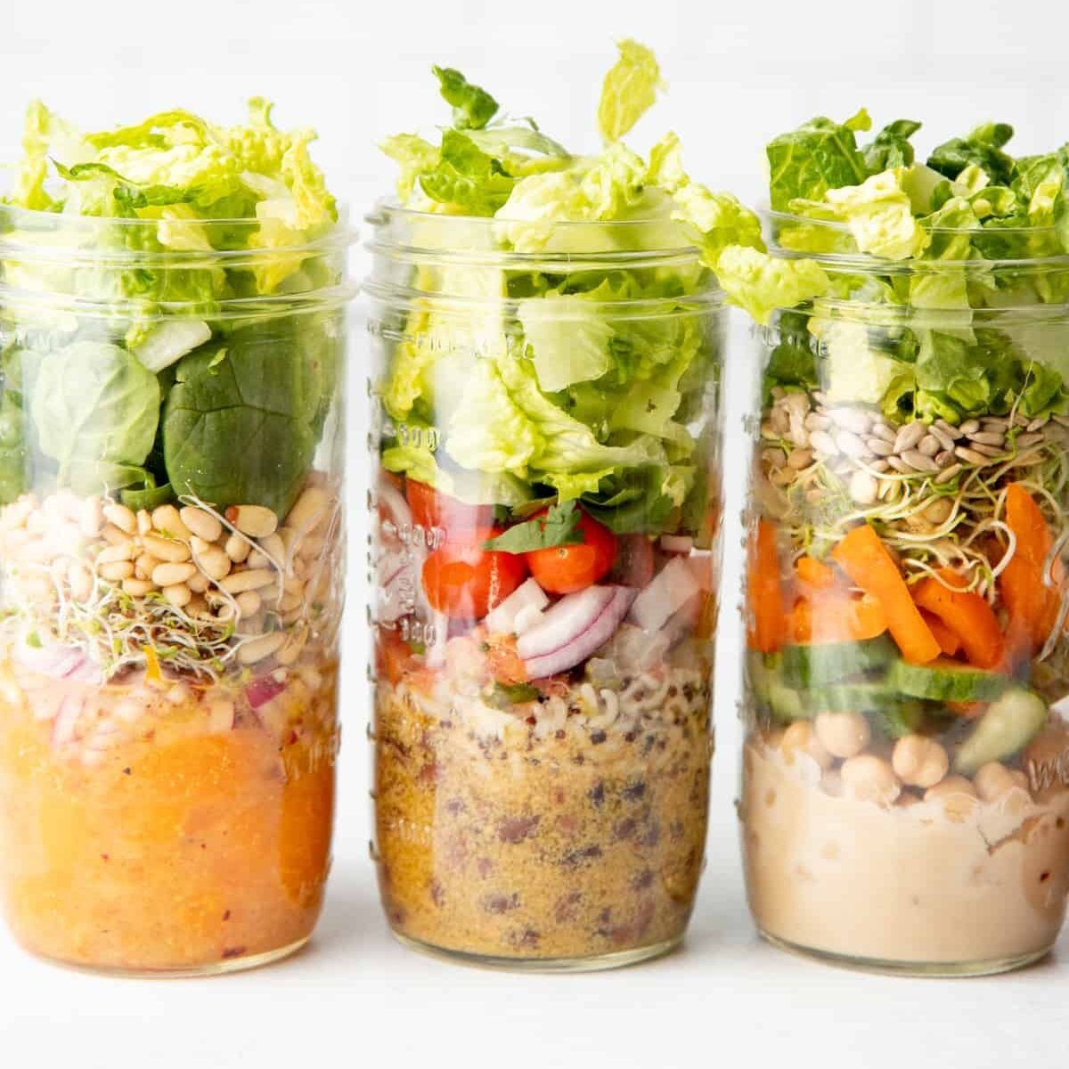 Three tall mason jars sit side-by-side in front of a white wall. Jars are layered with salad ingredients.