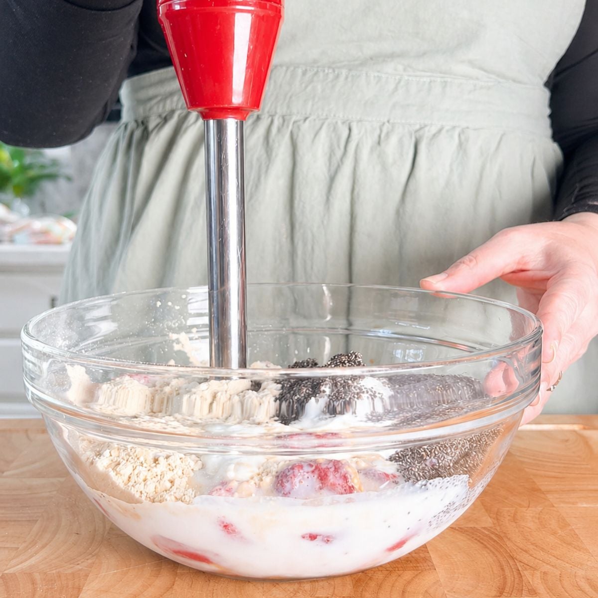 Immersion blender in a glass bowl with strawberries, milk, protein powder, and chia seeds before blending