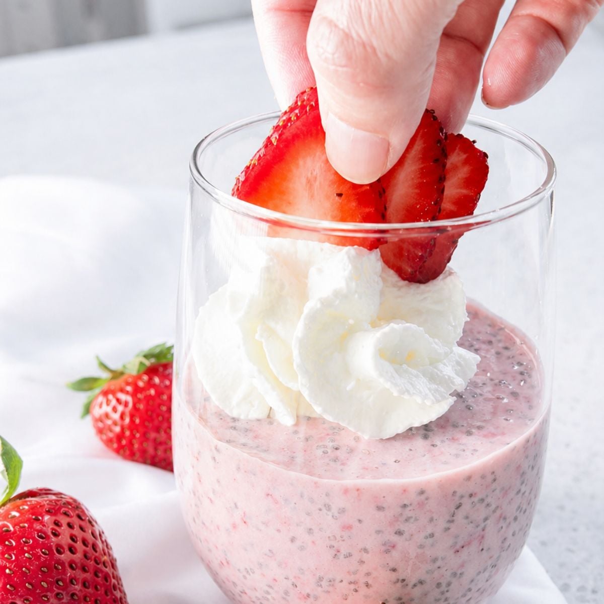 Hand placing a sliced fresh strawberry on top of a glass of strawberry chia pudding with whipped cream