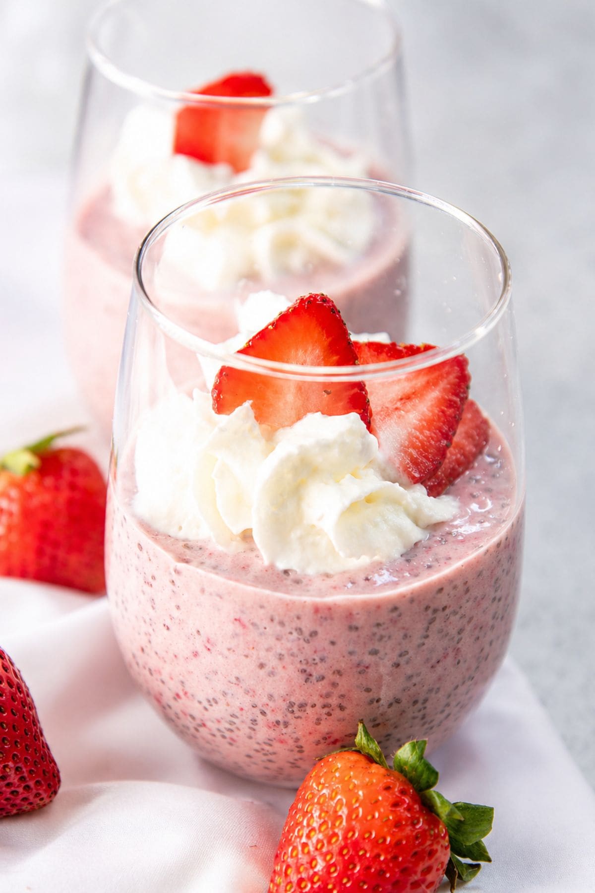 Two stemless wine glasses filled with creamy pink strawberry chia pudding, topped with whipped cream and fresh sliced strawberries