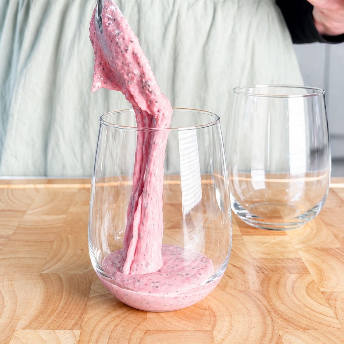 Thick pink strawberry chia pudding being poured from a spoon into a stemless wine glass