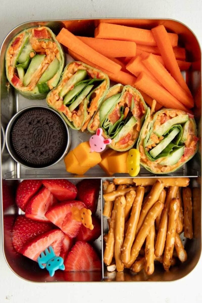 Overhead of bento-style lunchbox pack with veggie pinwheels, carrot sticks, pretzel sticks, sliced strawberries, cheese cubes, and an oreo cookie.